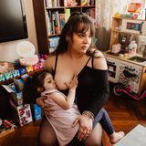 Eine Mutter hat ihr Kind auf dem Schoß und stillt es. Sie sitzt im Wohnzimmer, im Hintergrund steht eine Kinder-Spielküche