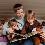 Eine Mutter sitzt mit ihren beiden Kindern auf dem Bett und liest ein Buch vor. Ein Kind nuckelt dabei an der der Brust