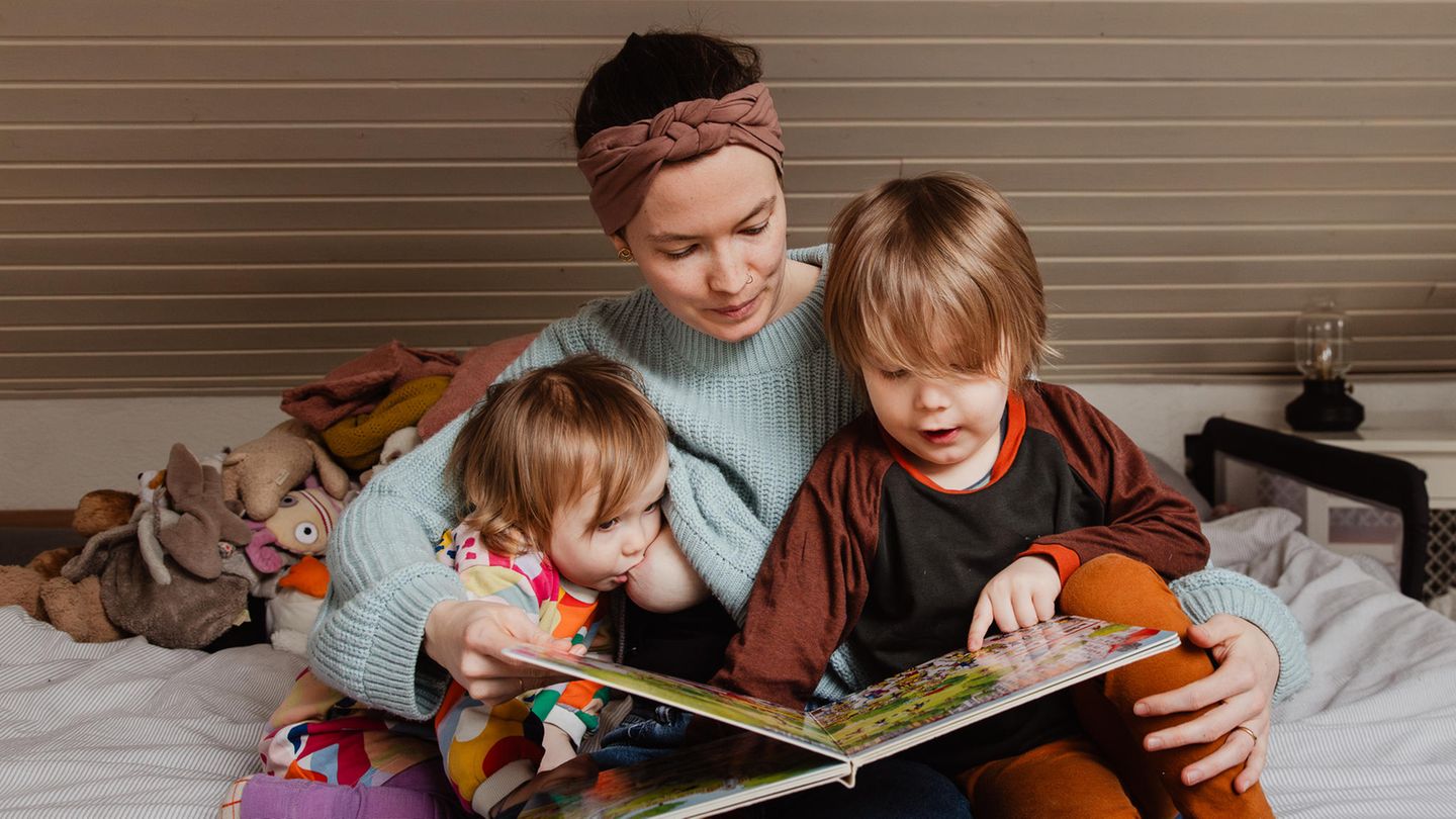 Eine Mutter sitzt mit ihren beiden Kindern auf dem Bett und liest ein Buch vor. Ein Kind nuckelt dabei an der der Brust