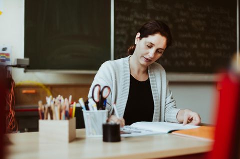 Lehrerin sitzt im Klassenzimmer am Schreibtisch und korrigiert im Schulheft – das macht sie auch in den Ferien