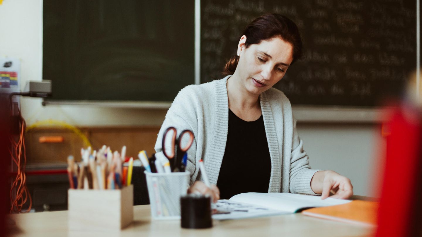 Lehrerin sitzt im Klassenzimmer am Schreibtisch und korrigiert im Schulheft – das macht sie auch in den Ferien