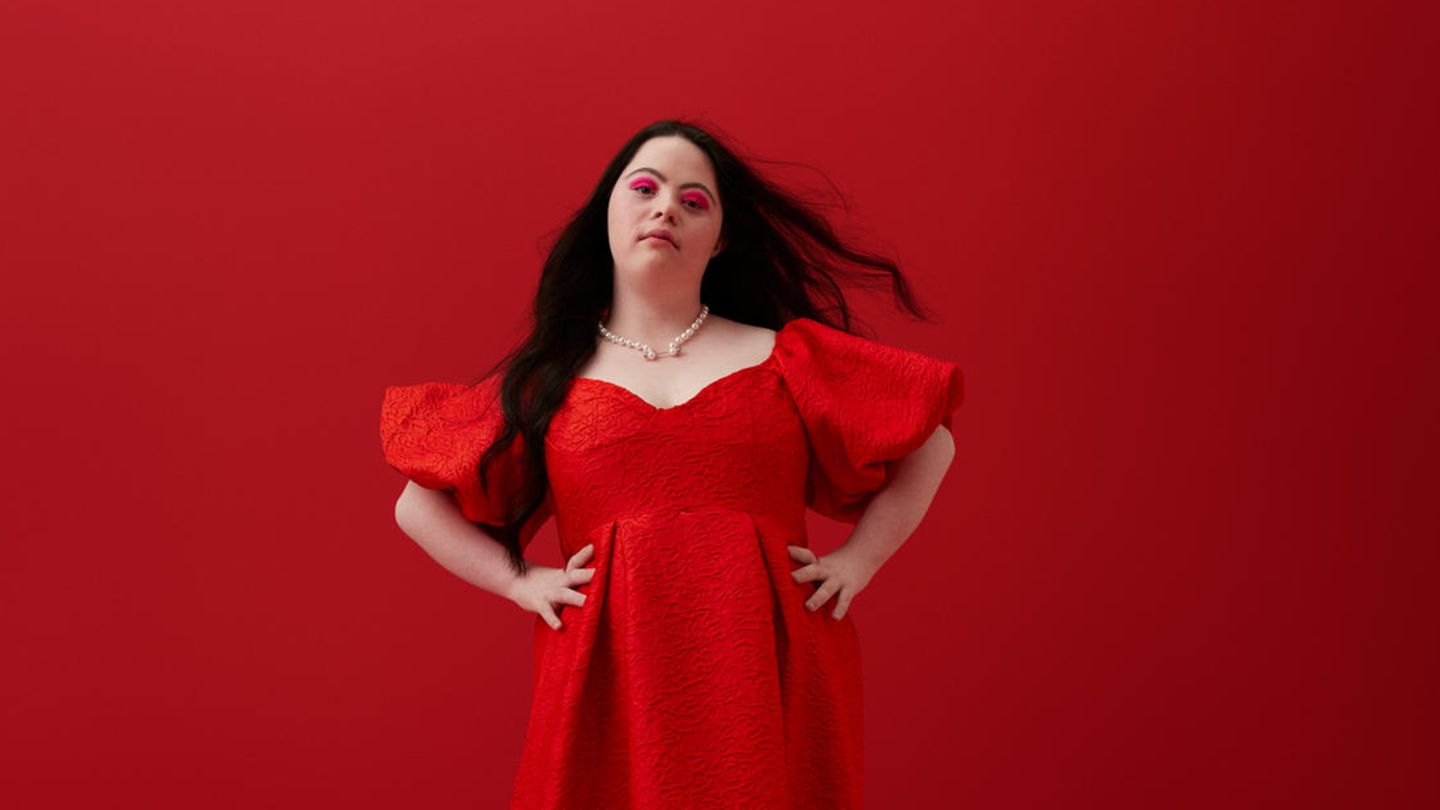 Eine Frau mit der Behinderung Trisomie 21 steht Selbstbewusst in einem roten Kleid vor einem roten Hintergrund