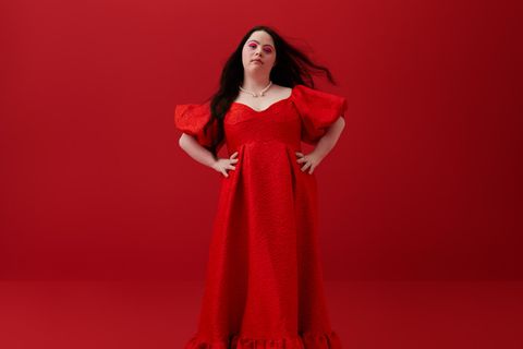 Eine Frau mit der Behinderung Trisomie 21 steht Selbstbewusst in einem roten Kleid vor einem roten Hintergrund