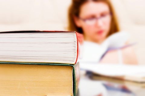 Im Vordergrund liegen zwei Bücher aufeinander. Im Hintergrund sitzt eine Frau mit Brille und liest