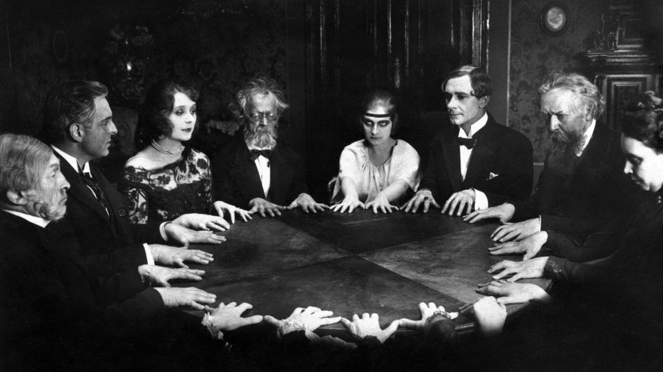 Schwarzweiß-Aufnahme von Menschen, die um einen Tisch sitzen und ihre Hände flach auf die Tischplatte gelegt haben