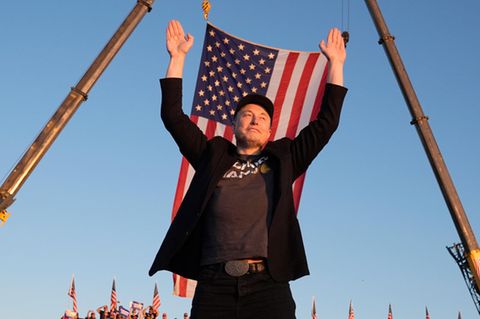 Elon Musk wirft die Arme hoch vor einer USA-Flagge