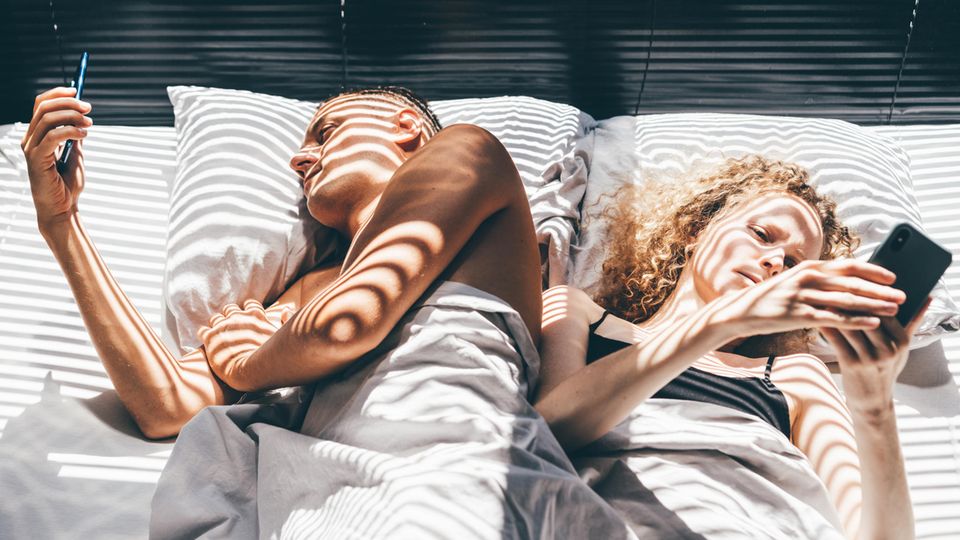 Symbolbild. Ein Pärchen liegt nebeneinander im Bett, beide sind an ihrem Handys.
