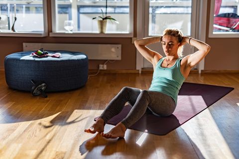 Eine Frau macht Sit-ups in ihrem Wohnzimmer. Sie liegt auf dem Boden und geht mit dem Oberkörper nach oben.