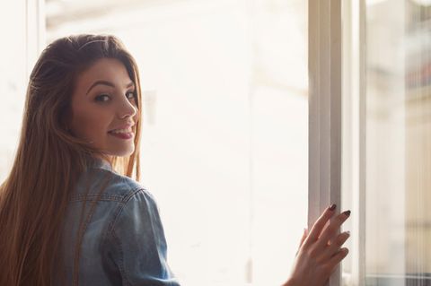 Eine Frau öffnet beide Fensterflügel zum Lüften und lächelt dabei über die rechte Schulter
