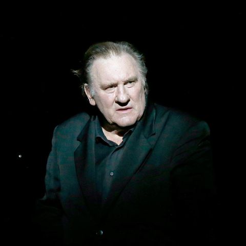 Gérard Depardieu steht in einem schwarzen Anzug von einem spotlight beleuchtet auf einer dunklen Bühne