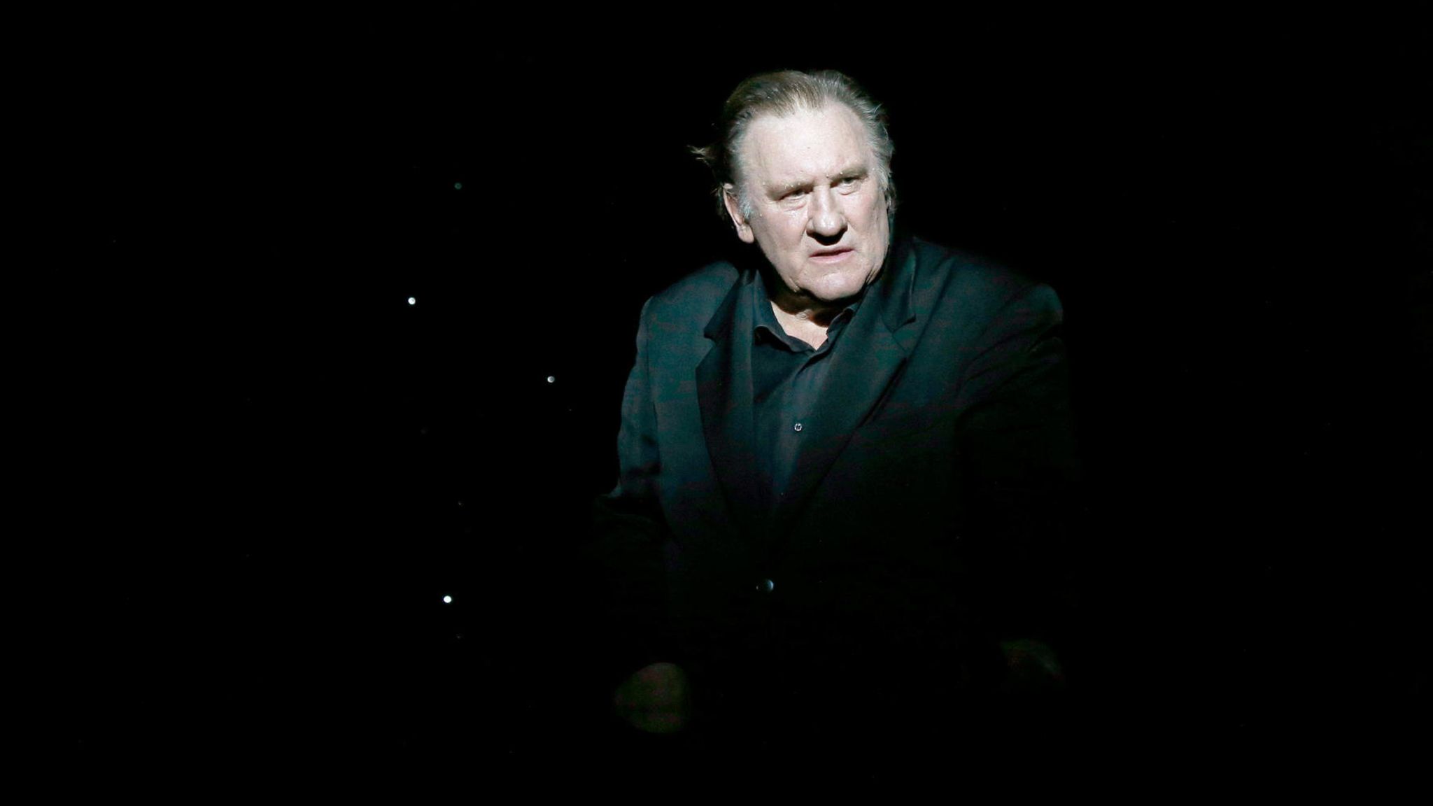 Gérard Depardieu steht in einem schwarzen Anzug von einem spotlight beleuchtet auf einer dunklen Bühne