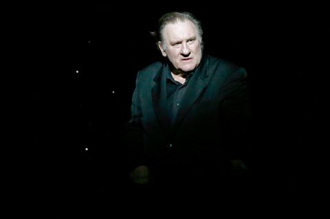 Gérard Depardieu steht in einem schwarzen Anzug von einem spotlight beleuchtet auf einer dunklen Bühne