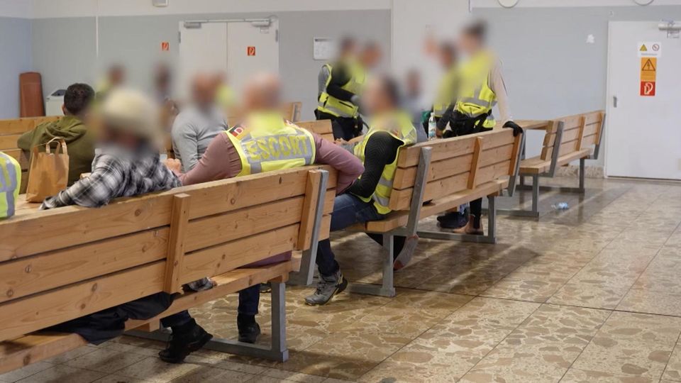 Polizisten in Warnwesten und Flüchtlinge sitzen in einer Wartehalle im Flughafen