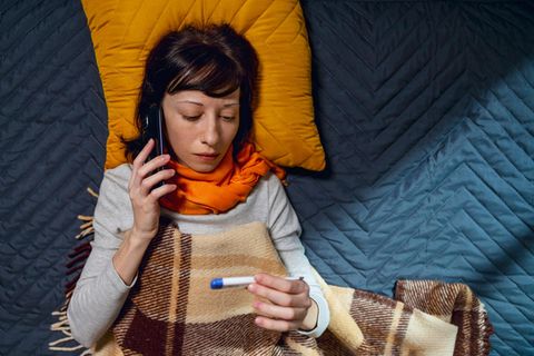Krankschreibung: Eine Frau liegt Krank im Bett und hält ein Thermometer in der einen und ein Handy am Ohr in der anderen Hand
