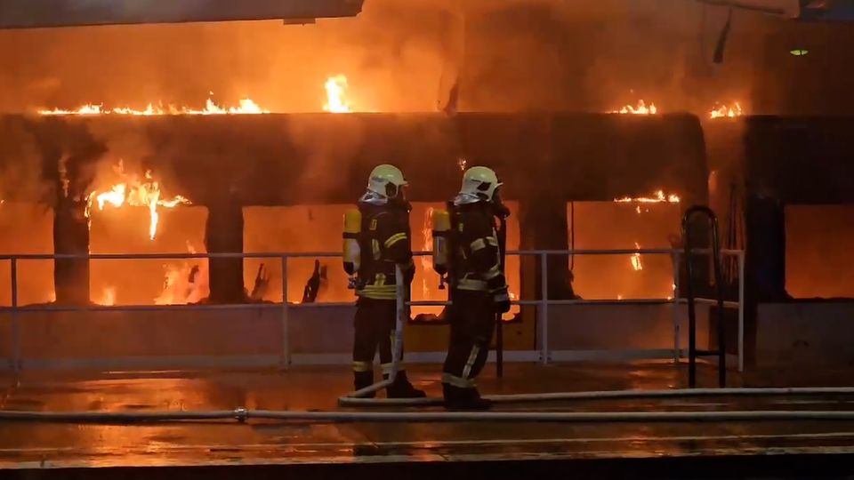 Zwei Feuerwehrleute stehen auf dem Bahnsteig in Berlin-Ahrensfelde und löschen einen brennenden Zug