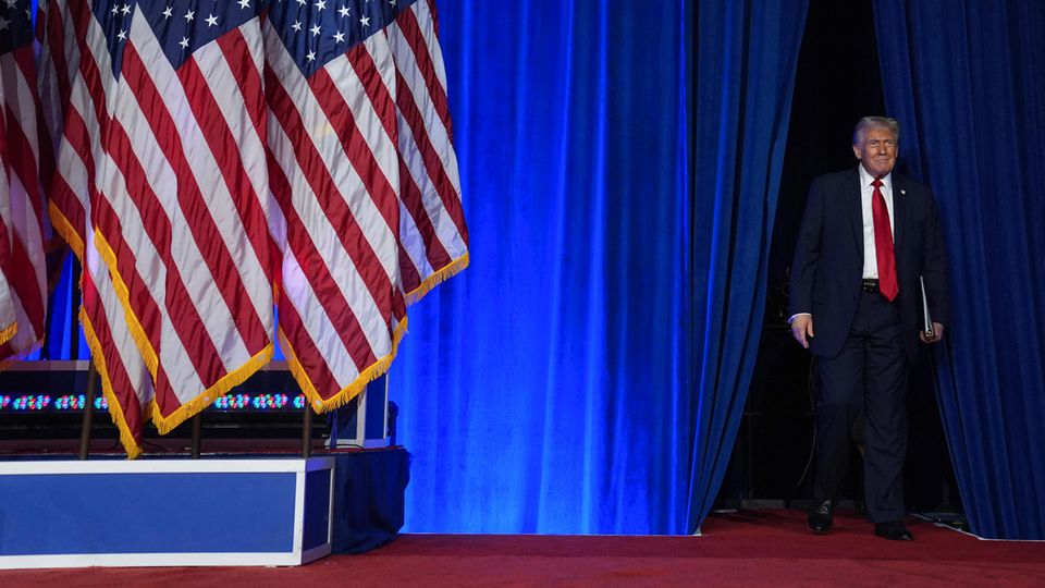 Trump, wie er durch einen blauen Vorhang, auf die Bühne läuft
