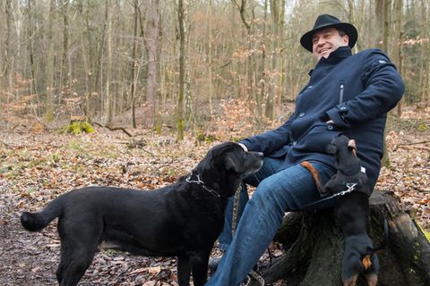 Markus Söder sitzt im Wald, vor ihm steht ein schwarzer Hund.