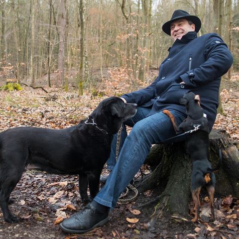 Markus Söder sitzt im Wald, vor ihm steht ein schwarzer Hund.