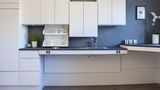 Die barrierefreie Küche  Wer seine Küche rollstuhlgerecht umgestalten möchte, kommt um Sonderlösungen nicht herum. Es gibt mittlerweile optisch attraktive Küchensysteme, deren Design nicht nach Krankenhaus aussieht. Der auf barrierefreie Küchen spezialisierte Hersteller Ropox bietet elektrisch höhenverstellbare Arbeitsplatten einschließlich Spülbecken, die sich mit dem Rollstuhl unterfahren lassen. Auch die ansonsten im Sitzen unerreichbare Hochschränke lassen sich elektrisch absenken. Das System ist auch praktisch, wenn man zusammen mit Kindern kochen oder backen möchte. Je nach Ausbaustufe liegen die Kosten zwischen 2000 und 4800 Euro. Einbauen kann es jedes Küchenstudio oder Schreiner.   Bei den Unterschränke gilt: ausfahrbare Schubladen statt Türen. Auf dem Kochfeld sollten die Induktionszonen nebeneinander liegen, damit im Sitzen nicht über heiße Töpfe gegriffen werden muss. Praktisch ist zudem ein flaches Spülbecken mit einer ausziehbaren Verlängerung des Wasserhahns. Der Backofen sollte zur Seite öffnen lassen und auf Höhe der Arbeitsplatte montiert sein.   Für die Gewährung von Fördermitteln über die Pflegestufe oder einen KfW-Kredit muss die DIN-Norm 18040-2 erfüllt werden. Es gibt Handwerksbetriebe, die neben Planung und Durchführung auch die Beantragung von Fördermitteln übernehmen. 