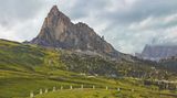 Eine Almwiese mit Gesteinsblöcken, Alpenpass-Straße und dem spitzen Bergmassiv des Monte Gusela
