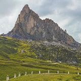 Eine Almwiese mit Gesteinsblöcken, Alpenpass-Straße und dem spitzen Bergmassiv des Monte Gusela