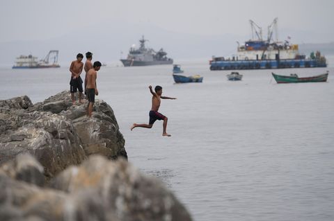 Jugendlicher springt von einer Klippe ins Meer, im Hintergrund mehrere große Schiffe
