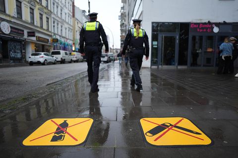 Zwei gelbe Piktogramme aufs Pflaster gemalt: Links eine zerbrochene Flasche, rechts ein Klappmesser – beide rot durchkreuzt