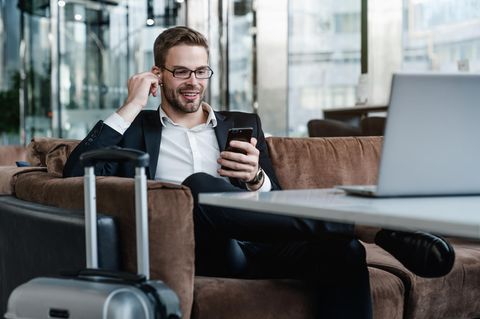 Mann mit AirPods, Handy und Gepäck telefoniert auf einem Sofa sitzend.