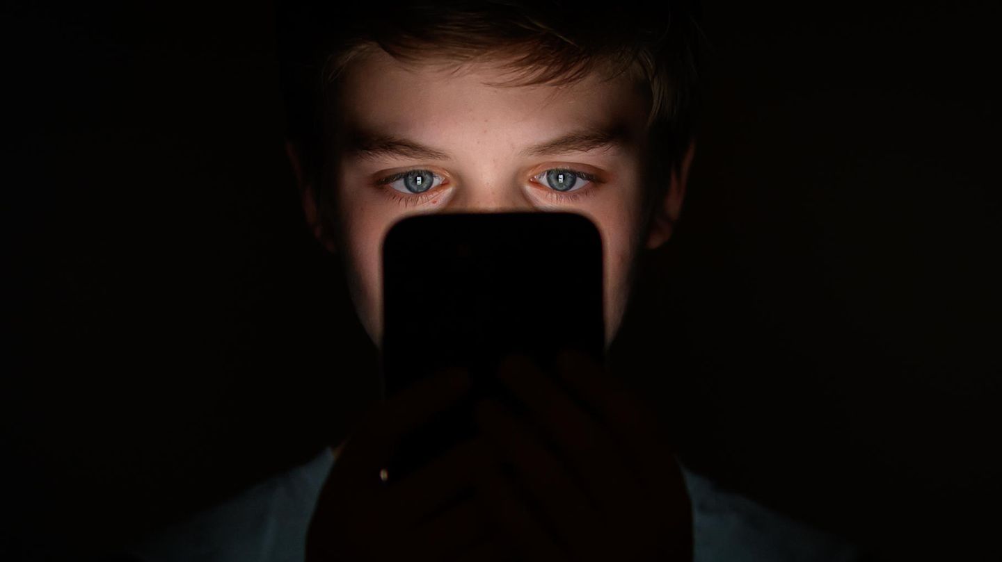Ein Junge sitzt im Dunkeln und schaut in den hellen Bildschirm seines Handys und scrollt auf Social Media