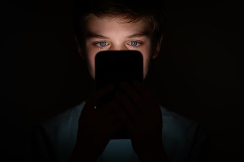 Ein Junge sitzt im Dunkeln und schaut in den hellen Bildschirm seines Handys und scrollt auf Social Media