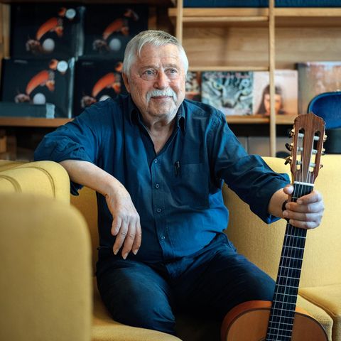 Wolf Biermann sitzt auf einer gelben Couch und hält eine Gitarre in der linken Hand