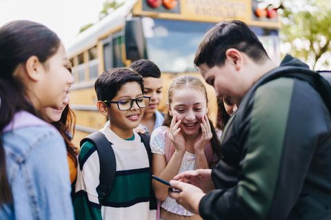 Schüler stehen vor einem Schulbus und gucken alle auf ein Handy und lachen