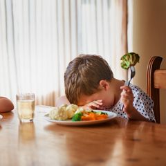 Zwei verzweifelte kleine Jungs sitzen jeder vor einem Teller mit Gemüse. Die gesunde Ernährung ist schwer