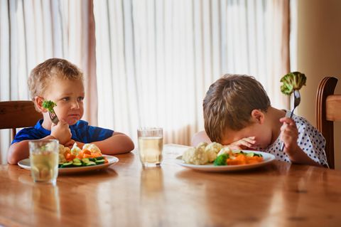 Zwei verzweifelte kleine Jungs sitzen jeder vor einem Teller mit Gemüse. Die gesunde Ernährung ist schwer