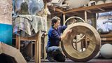 Eine kurzhaarige Frau schmirgelt eine große Holzscheibe, die ein Globus-Sockel wird