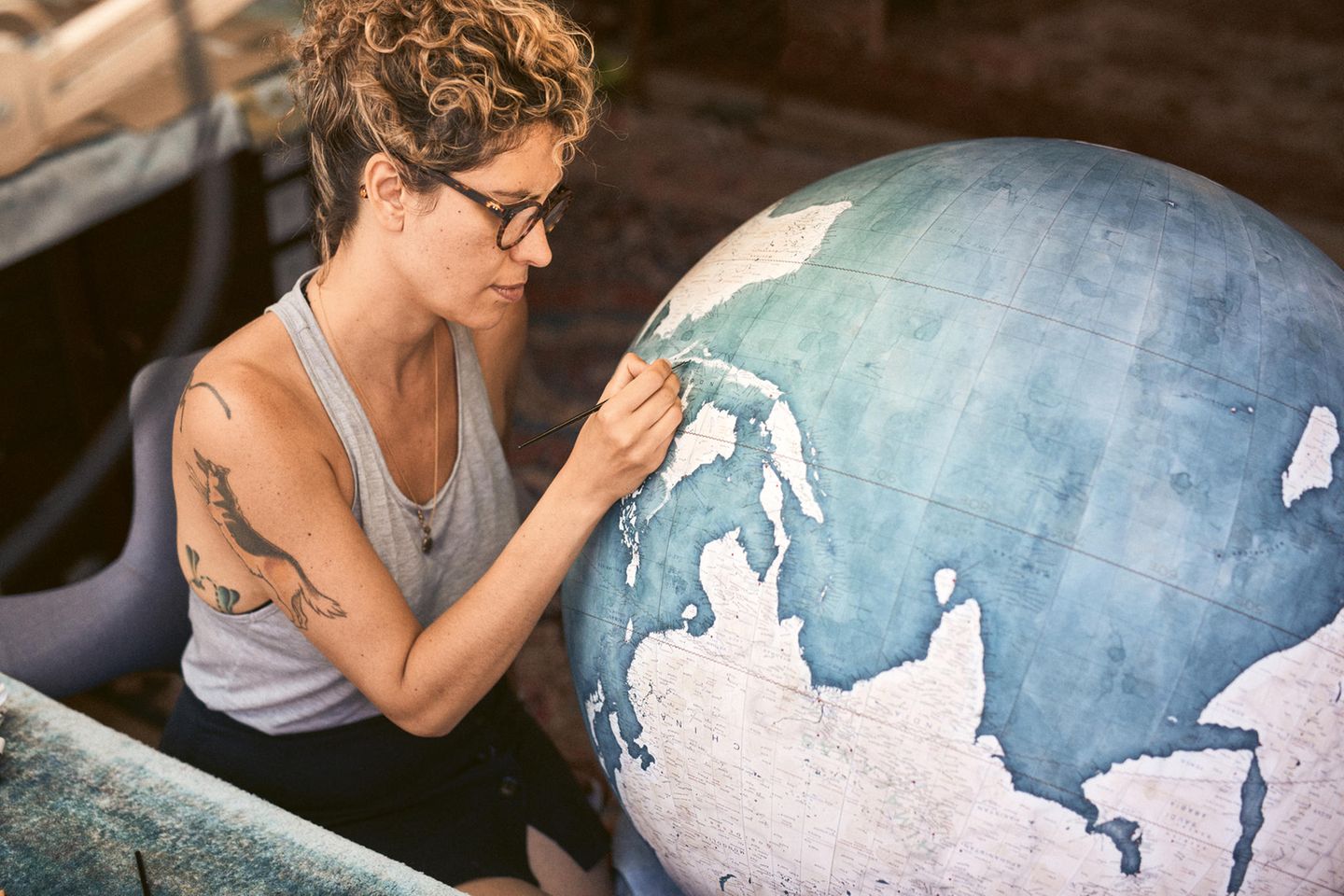 Eine junge Frau mit hochgesteckt Haaren und Brille bemalt einen Globus mit einem feinen Pinse