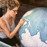 Eine junge Frau mit hochgesteckt Haaren und Brille bemalt einen Globus mit einem feinen Pinse