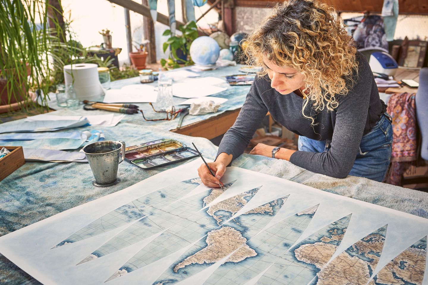 In der Globus-Werkstatt bemalt eine junge Frau mit Locken mit einem Pinsel eine Landkarte am Tisch.