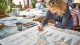 In der Globus-Werkstatt bemalt eine junge Frau mit Locken mit einem Pinsel eine Landkarte am Tisch.