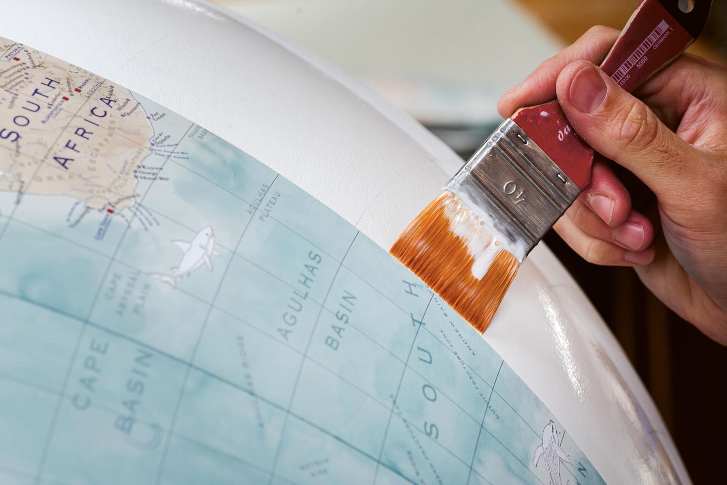 Auf eine weiße Gipskugel wird mit einem Pinsel Kleber aufgetragen, um die Globus-Landkartenstreifen aufzukleben