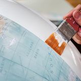 Auf eine weiße Gipskugel wird mit einem Pinsel Kleber aufgetragen, um die Globus-Landkartenstreifen aufzukleben