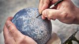 Zwei Hände an einem handballgroßen Mond-Globus, die eine hält, die andere bemalt ihn mit einem Stift