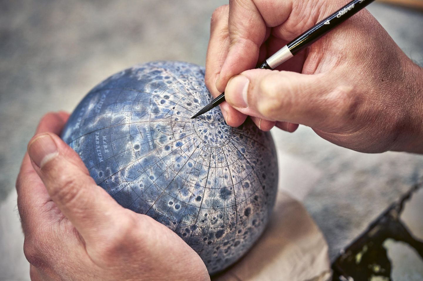 Zwei Hände an einem handballgroßen Mond-Globus, die eine hält, die andere bemalt ihn mit einem Stift