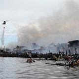 Ein Hubschrauber nährt sich mit Wasser zum Löschen, im Wasser vor dem Brand sind Bewohner mit ihren Sachen zu sehen