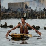 Eine Person auf ihrem kleinen selbstgezimmertem Floß von 1 Quadratmeter, in den Händen Holzbretter als Paddel