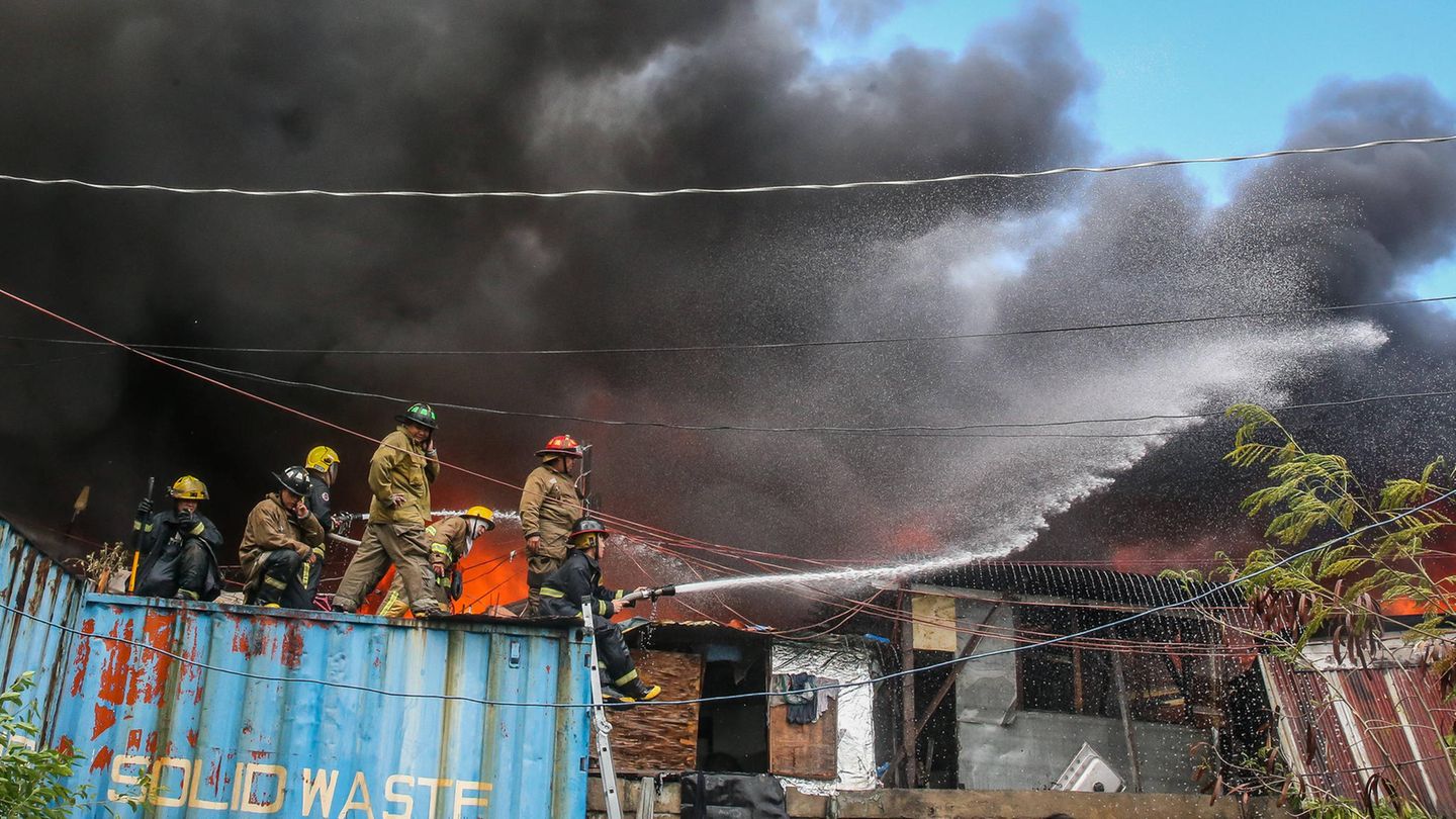 Fünf Feuerwehrleute stehen auf einem Container und sprühen Wasser auf die brennenden Häuser