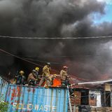 Fünf Feuerwehrleute stehen auf einem Container und sprühen Wasser auf die brennenden Häuser