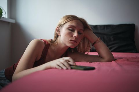 Dating-Krise: Frau lehnt am Bett, Handy in der Hand