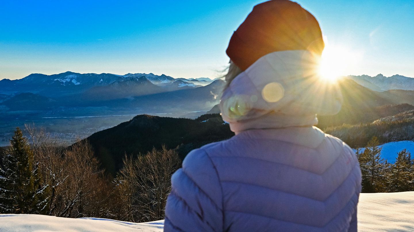 Eine Frau in dicker Jacke schaut bei Sonnenschein über Berge mit Schnee