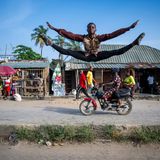 Ein junger Tänzer springt auf einer Straße, auf der ein Motorrad fährt, im Spagat.